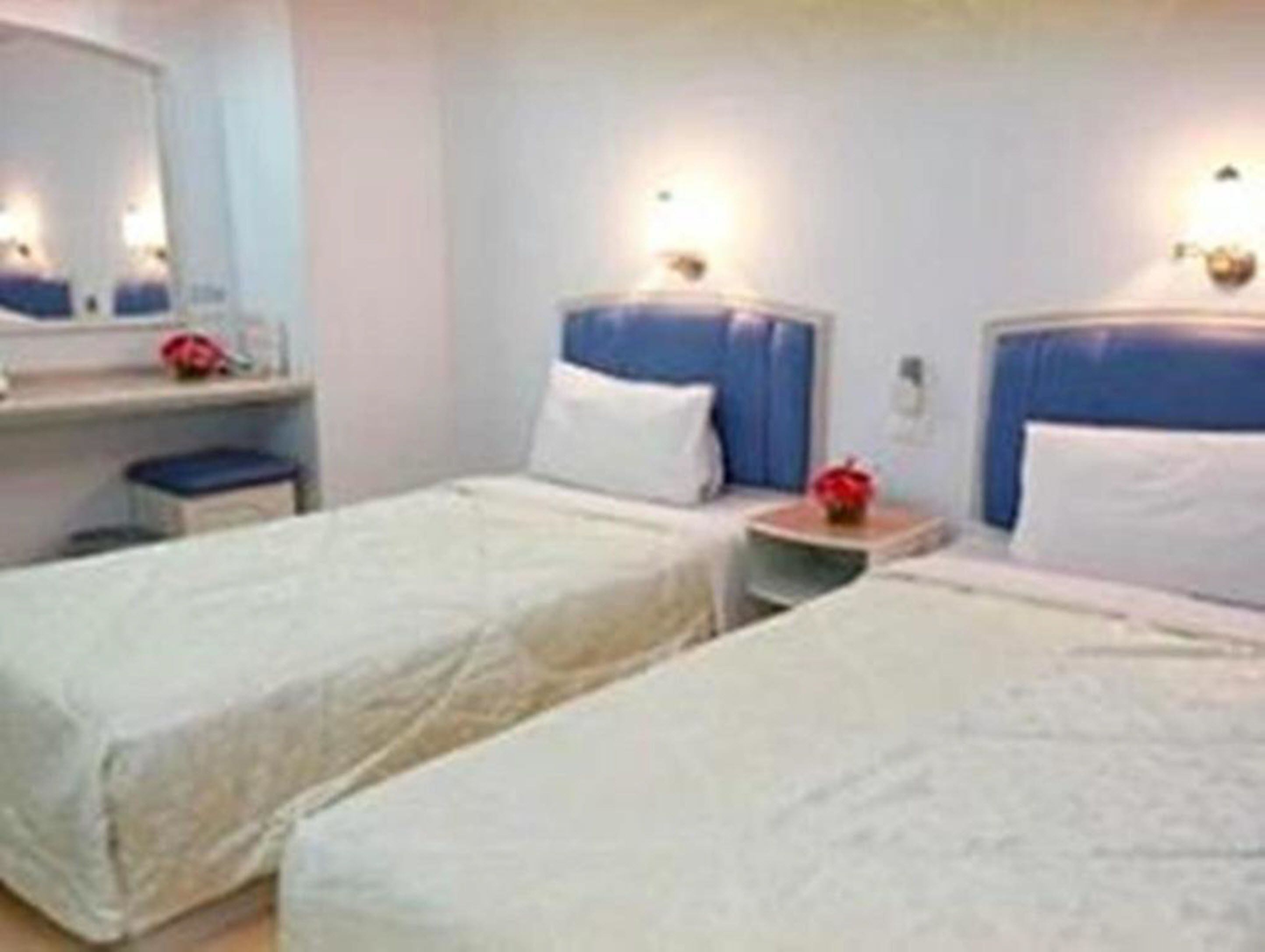 Habitación de hotel con dos camas individuales, una mesita de noche pequeña con lámpara, un espejo y una mesita con una flor roja.