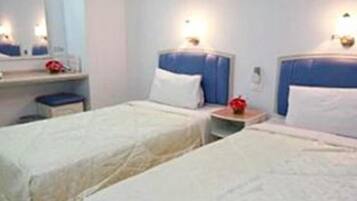 Habitación de hotel con dos camas individuales, una mesita de noche pequeña con lámpara, un espejo y una mesita con una flor roja.