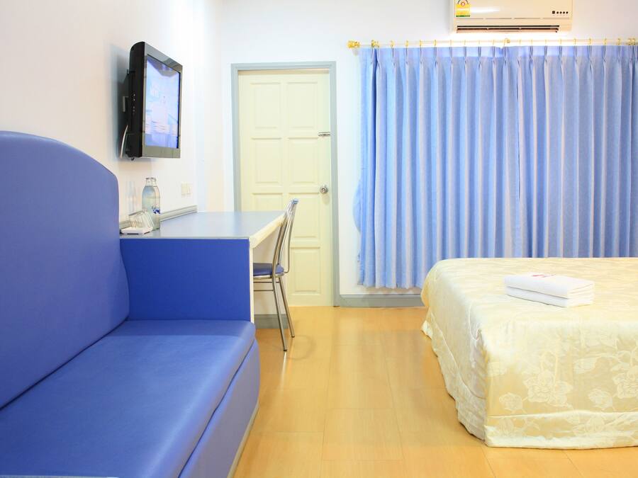 A hotel room with a blue sofa, a bed with a floral bedspread, a television mounted on the wall, and a window with blue curtains.