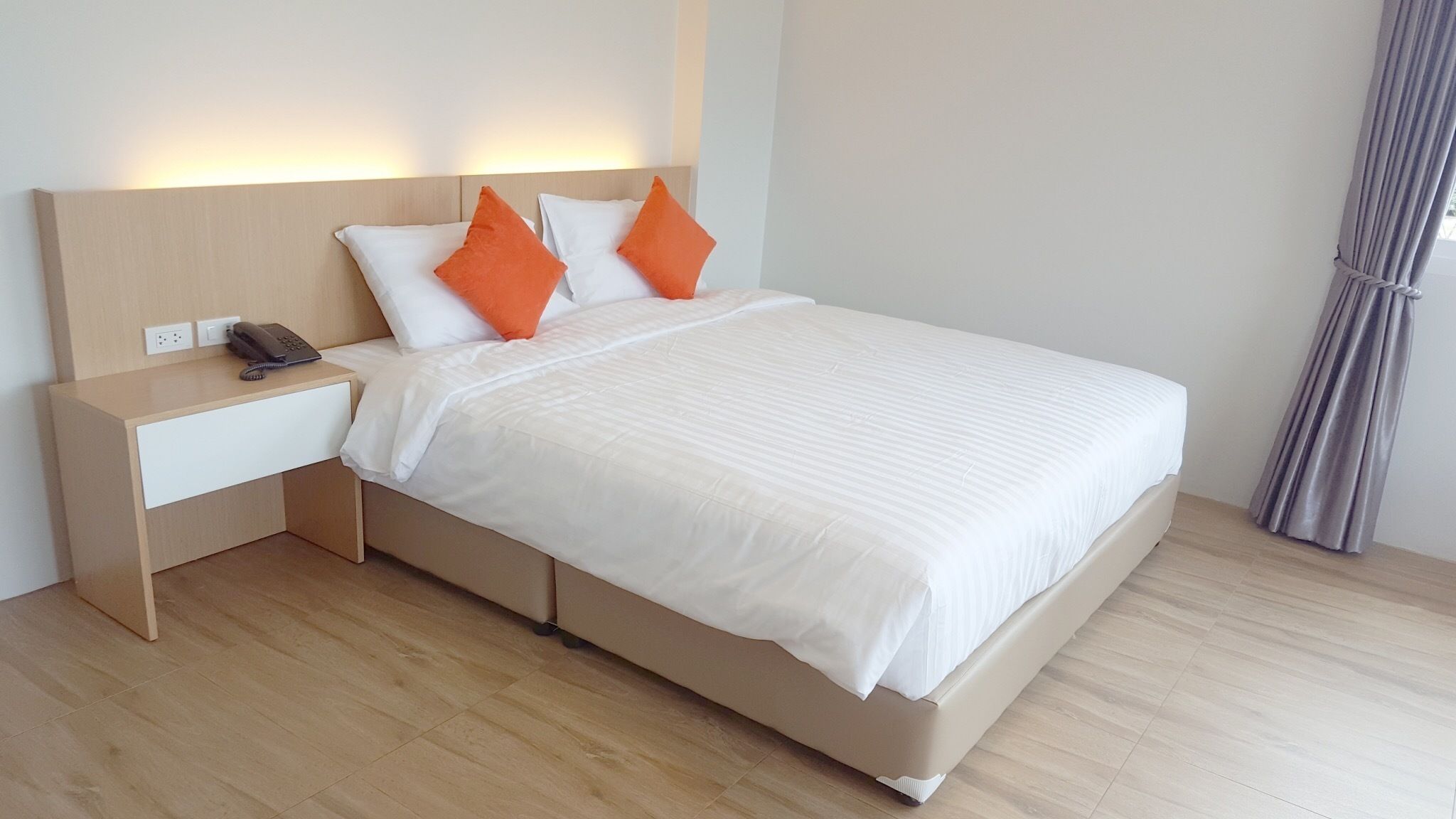A modern hotel room with a large bed, white bedding, two orange pillows, a bedside table with a phone, and a nightstand.