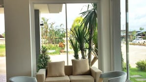 A modern living room with a beige sofa, two grey armchairs, and a small round table. Large windows offer a view of a garden and a street. - Sleeping Tree Hotel (Fang)