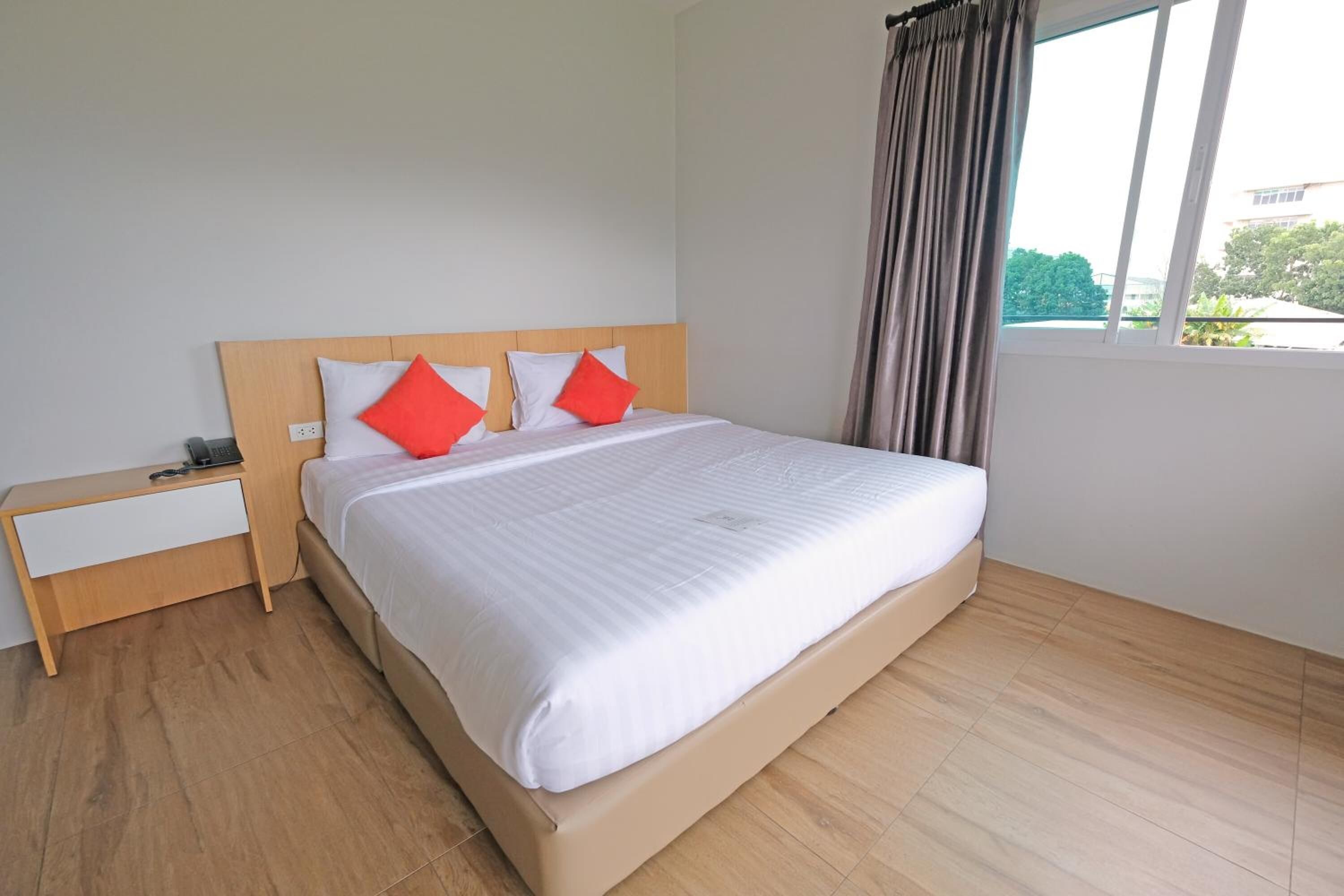 A neatly made bed with white bedding and two red pillows, a wooden nightstand with a phone, and a window with curtains.