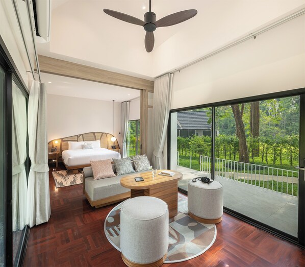 A modern bedroom with a large bed, a sofa, a coffee table, and a balcony with a view of greenery.