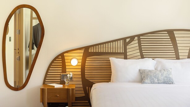 Habitación de hotel con cama, mesita de noche con lámpara, espejo y cabecero de madera.