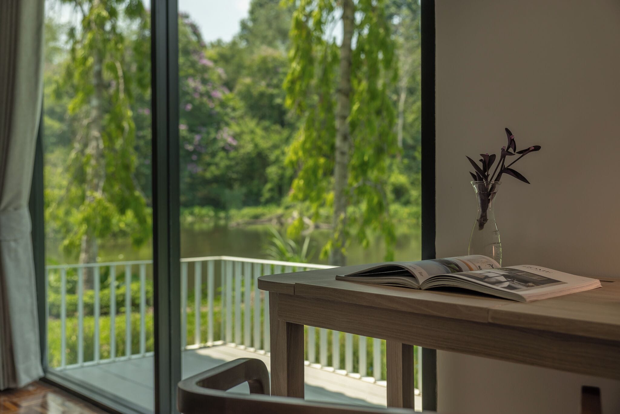 A room with a large window, a wooden table with a vase and magazine, and a view of greenery outside.