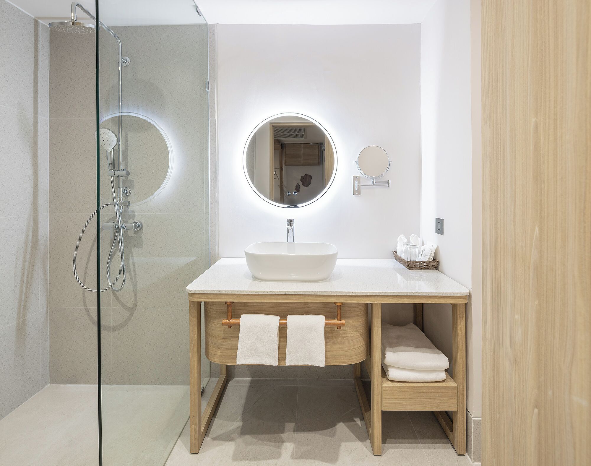 Un baño moderno con ducha envuelta en vidrio, espejo redondo y tocador de madera con lavamanos y perchero.