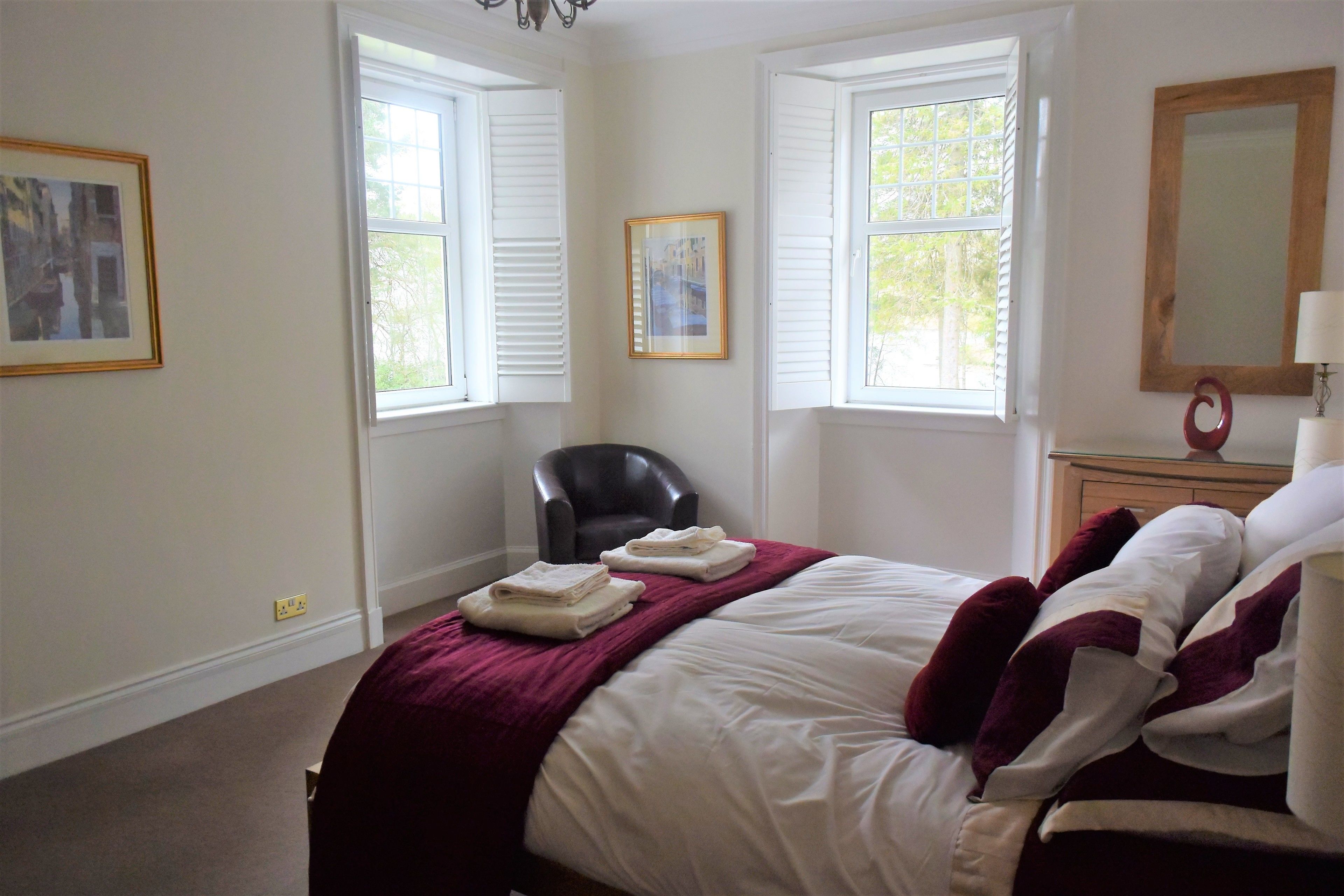 A hotel room with a bed, two windows with shutters, a chair, a bedside table with a lamp, and a mirror.