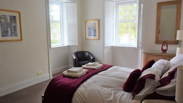 A hotel room with a bed, two windows with shutters, a chair, a bedside table with a lamp, and a mirror.