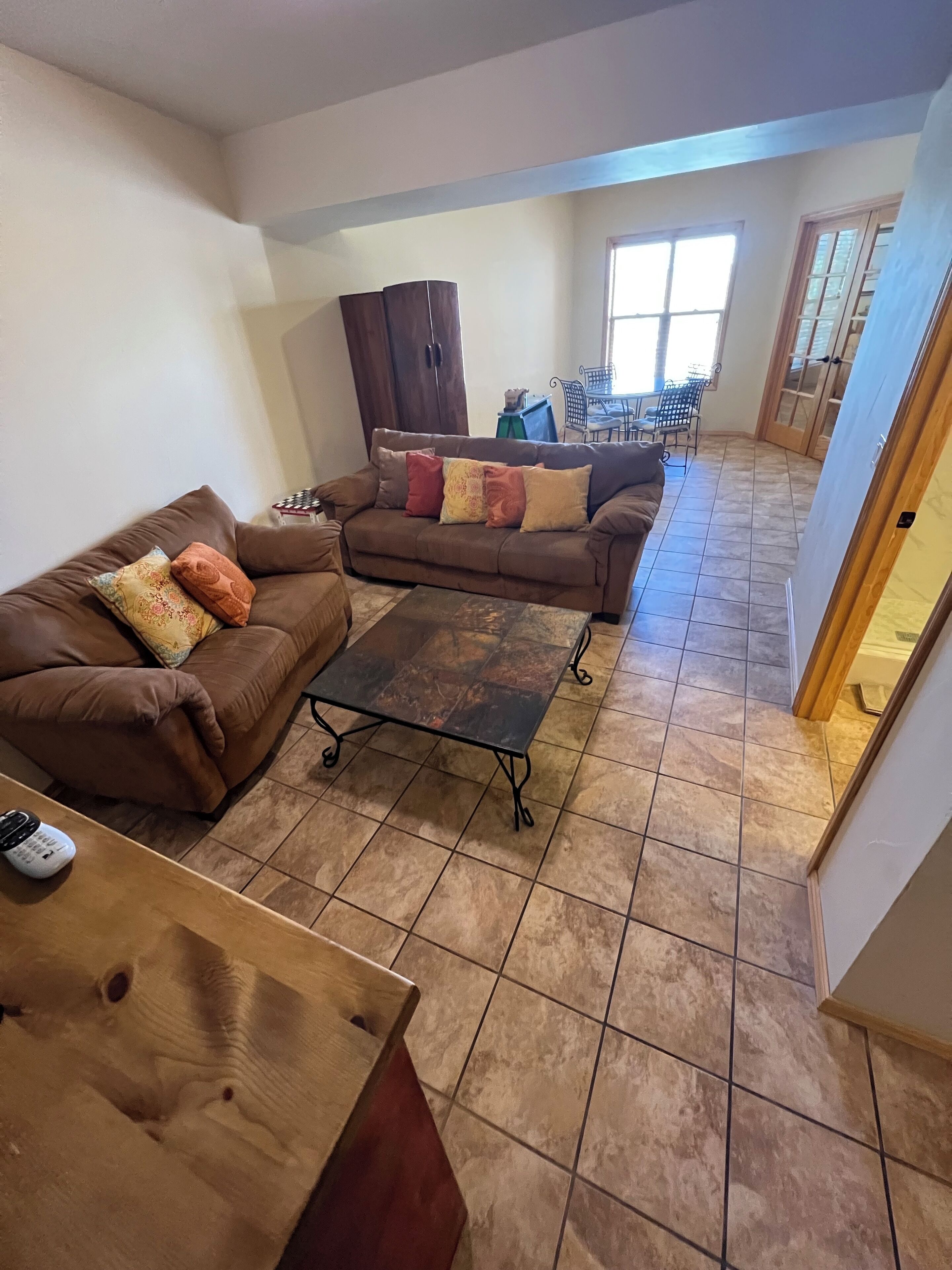 A living room with a brown sofa, a coffee table, and a wooden table. There is a window with a view of the outside.
