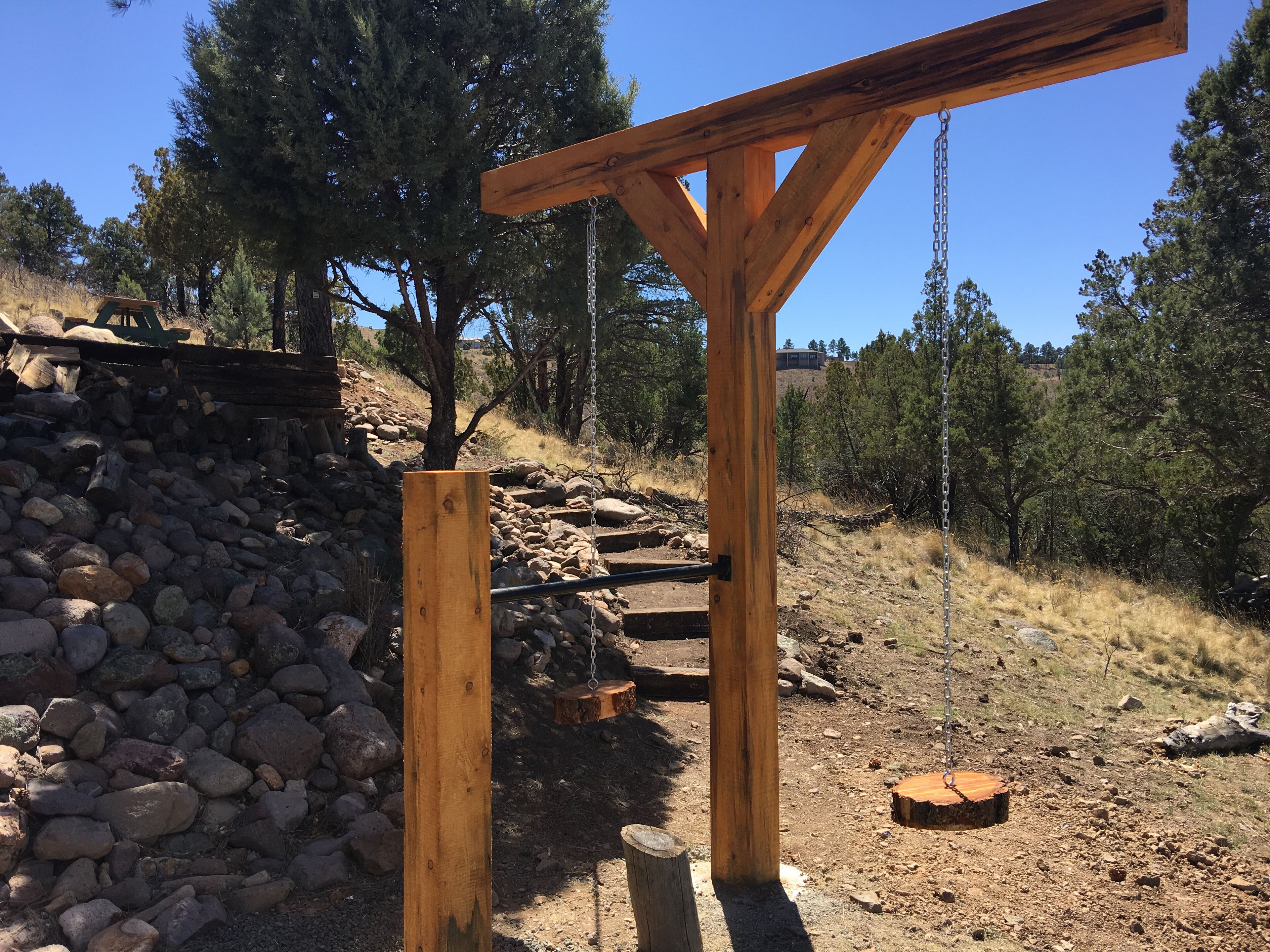A wooden pergola structure with a chain swing, surrounded by a rocky terrain and trees.