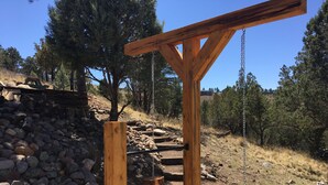 A wooden pergola structure with a chain swing, surrounded by a rocky terrain and trees.