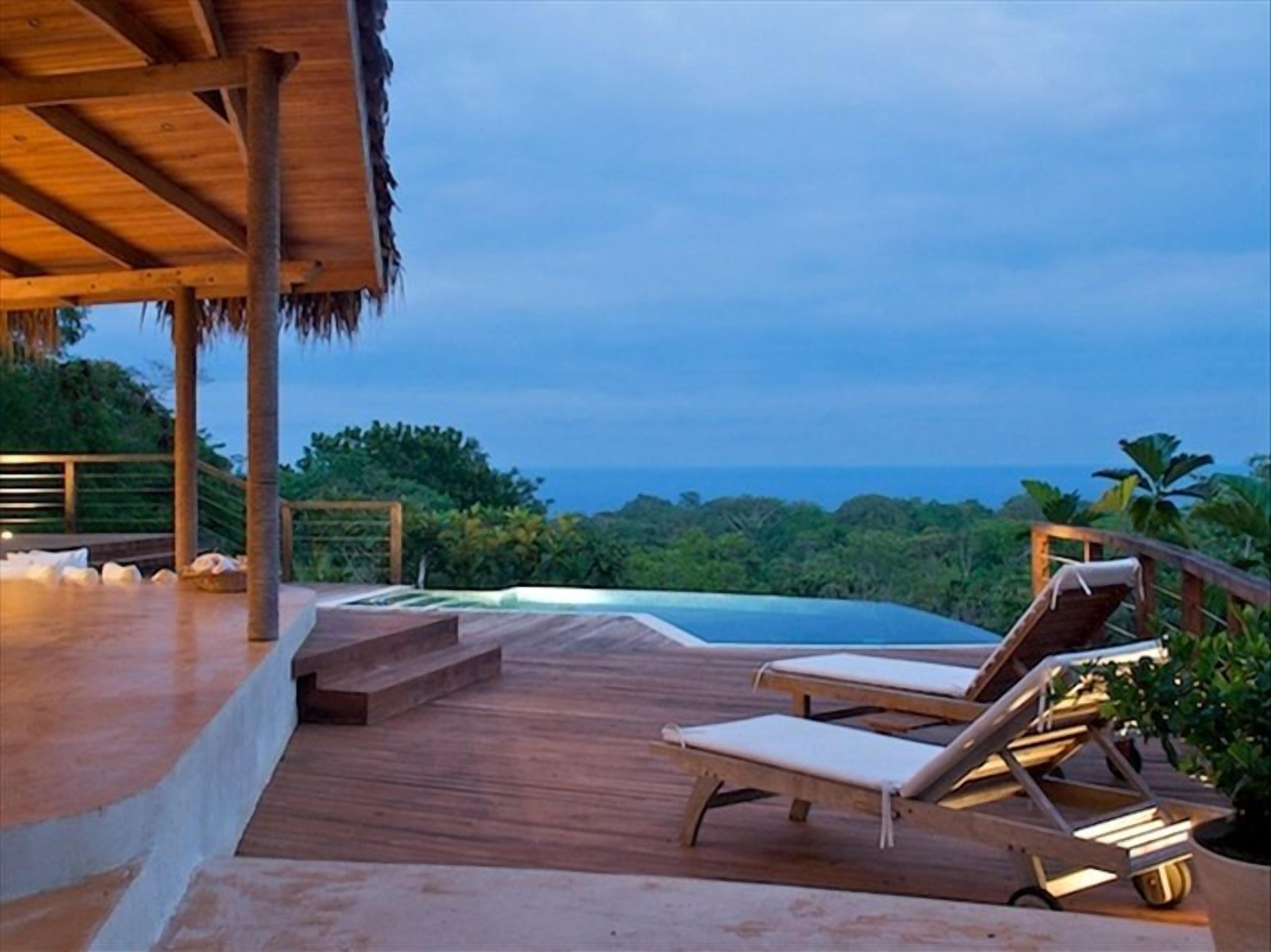 A wooden deck with lounge chairs and a small pool, overlooking a lush landscape and the ocean.