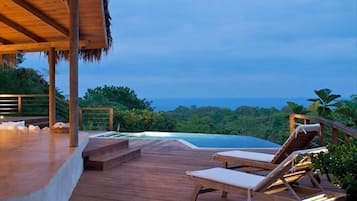 A wooden deck with lounge chairs and a small pool, overlooking a lush landscape and the ocean.