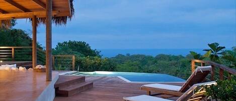 Una terraza de madera con sillones de descanso y una piscina pequeña, con vistas a un paisaje exuberante y el océano.
