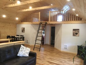 A spacious loft with a wooden floor, a black leather sofa, a dining table with chairs, a ladder to the upper level, and a staircase.