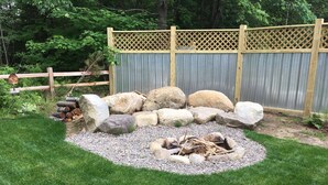 Un jardín con un pozo de fuego rodeado de rocas y troncos, una cerca de madera y un césped.