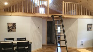 Una habitación con piso de madera, una mesa de comedor con sillas, una escalera y una escalera que sube a una zona superior.