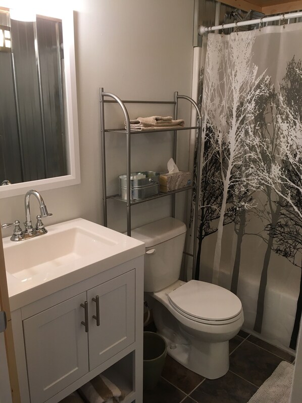 A bathroom with a white sink, a toilet, a metal shelf unit with toiletries, and a shower curtain with a tree design.