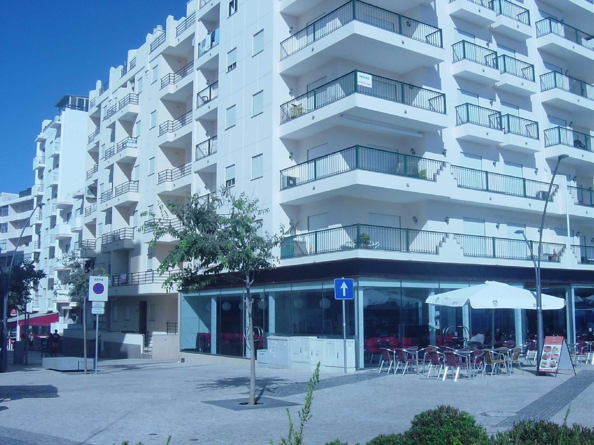 Ein mehrgeschossiges Wohnhaus mit Balkonen, ein Café mit Außenbestuhlung und einem strahlend blauen Himmel.