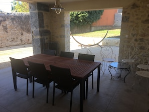 Un espace extérieur abrité, aménagé pour les repas, avec une table et des chaises en bois, un barbecue en métal et une vue sur le jardin.
