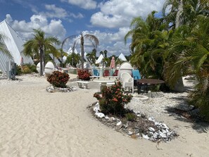 Un resort frente al mar con chalets blancos, palmeras y una zona de arena con asientos y una piscina pequeña.