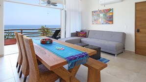 A spacious living area with a wooden dining table, grey sofa, and a view of the ocean.