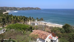 A coastal view with a beach, palm trees, and a building with a tiled roof.