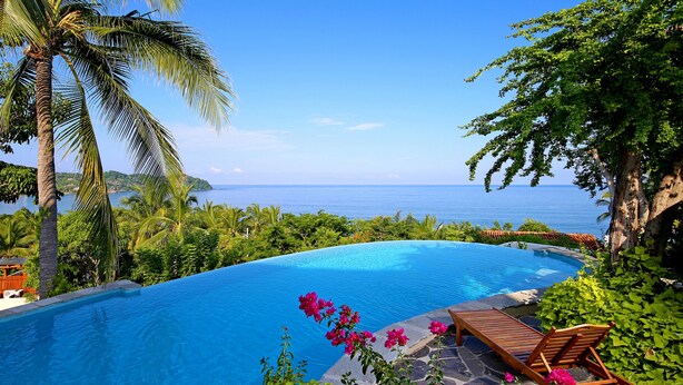 A swimming pool with a view of the ocean, surrounded by lush greenery and a wooden lounge chair.