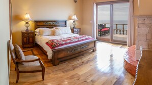 Quarto com cama de madeira, duas mesas de cabeceira com abajures, uma cadeira e vista para o oceano através de uma porta de correr de vidro.