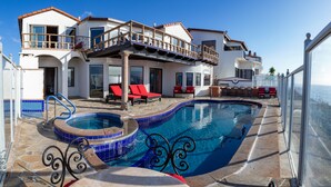 Piscina com deck de pedra e espreguiçadeiras vermelhas, cercada por uma casa de dois andares com varandas e um céu azul límpido.
