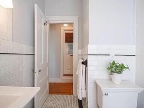 A bathroom with a white bathtub, a toilet, and a shower area. There is a towel hanging on the wall and a potted plant on the counter. - Ocean View East End Studio in a Historic Victorian (Portland)