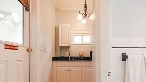 A narrow hallway leading to a kitchen with white cabinets, a black countertop, and a hanging light fixture. - Ocean View East End Studio in a Historic Victorian (Portland)