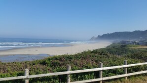 A coastal view with a sandy beach, a wooden fence, and a forested hill in the background.
