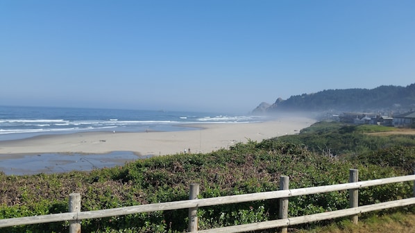 Vista costera con una playa arenosa, una valla de madera y una colina boscosa al fondo.