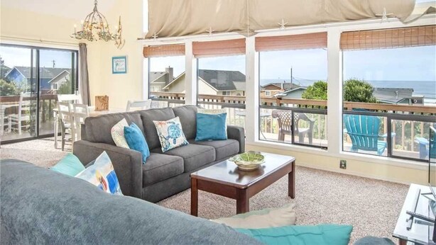 A spacious living room with a gray sofa, blue cushions, and a wooden coffee table. Large windows offer a view of the ocean and neighboring houses.