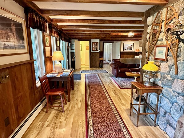 A cozy living room with a stone wall, wooden beams, a fireplace, and a dining area with a long red rug.
