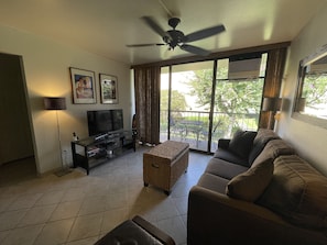 Un salon avec un canapé, un téléviseur posé sur une table en verre, un ventilateur de plafond et un balcon avec une table et des chaises.