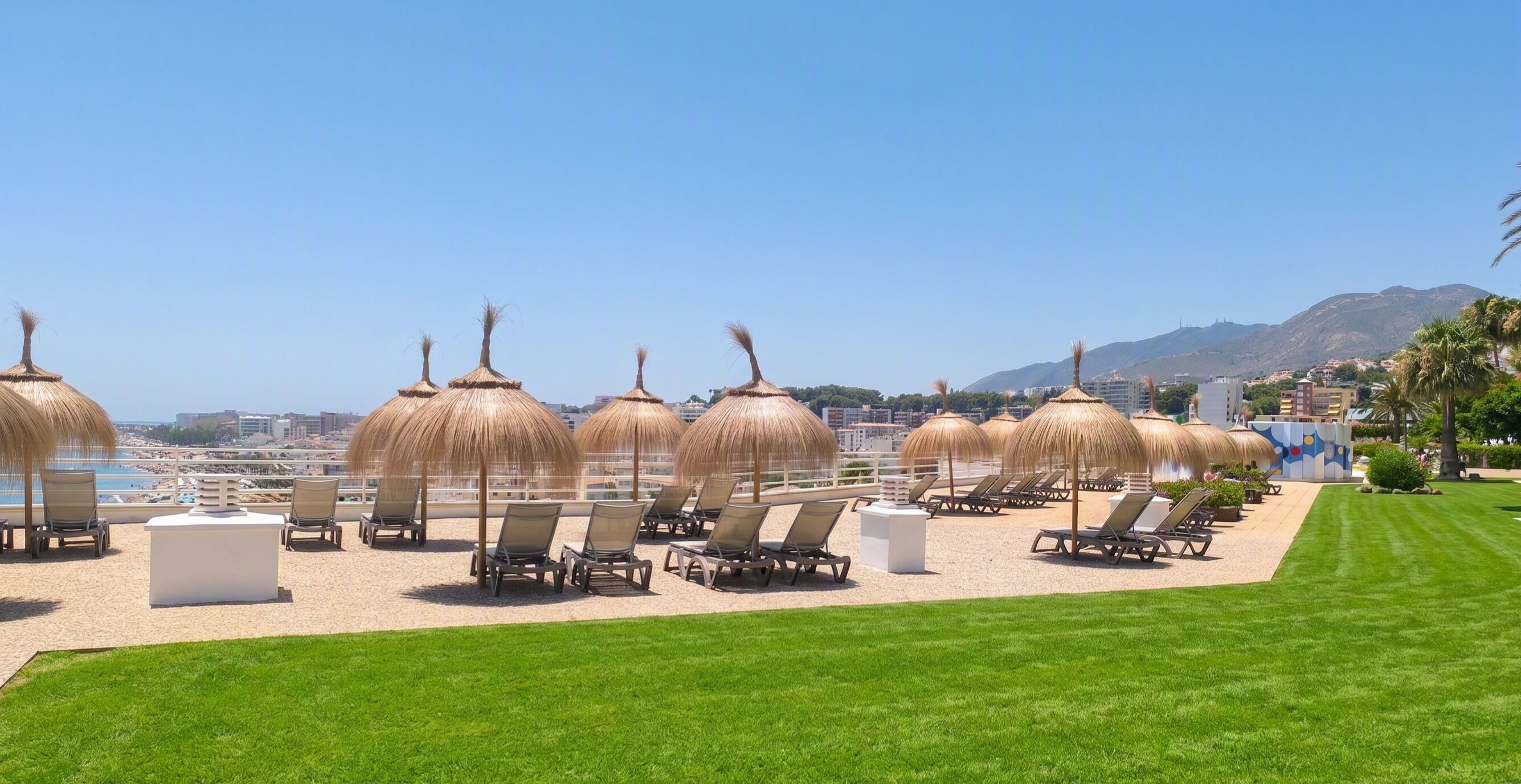 A beachside lounge area with sun umbrellas and lounge chairs, a grassy lawn, and a view of the beach and mountains.