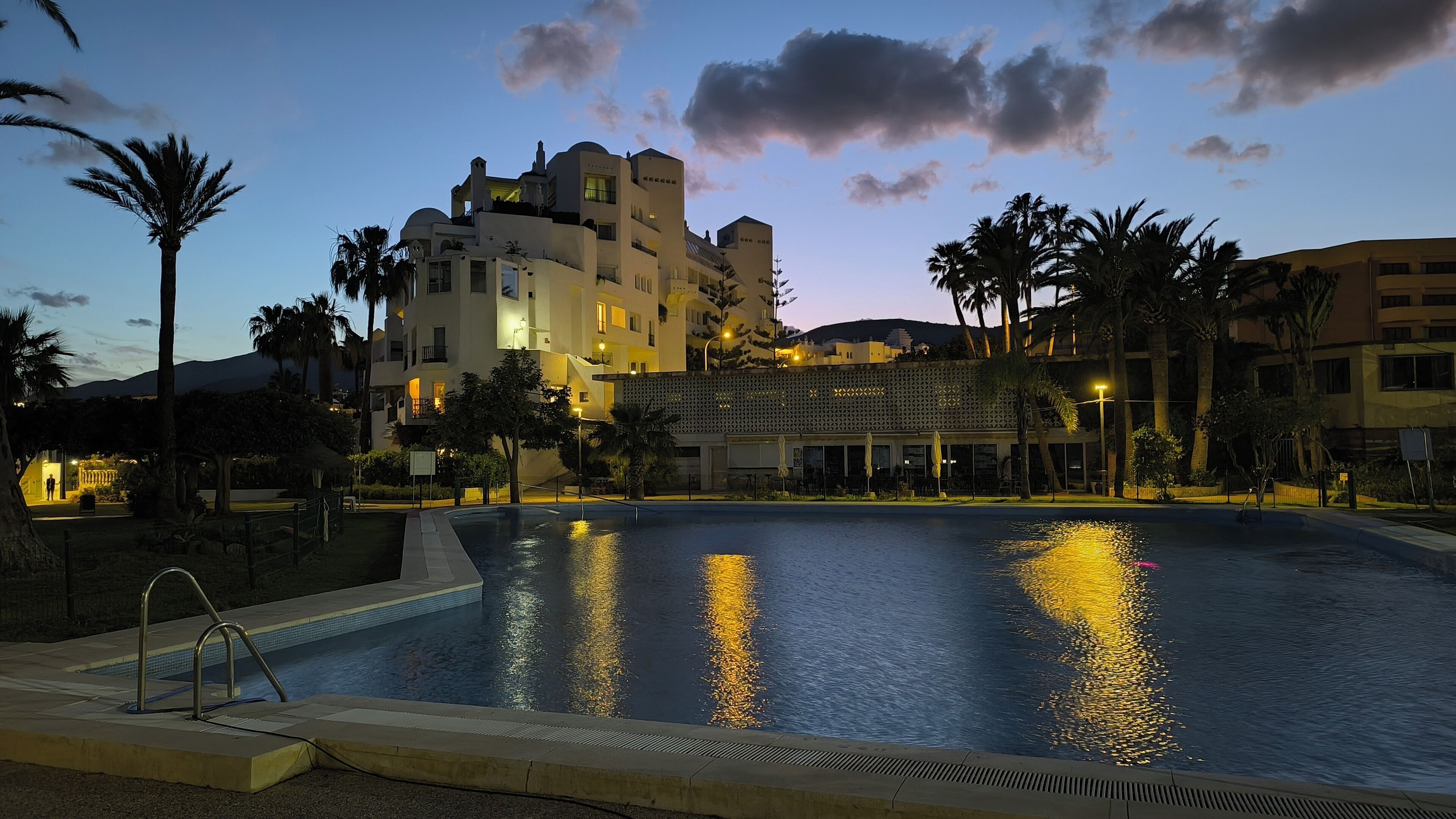 Une piscine dont la surface reflète les lumières, entourée de palmiers et bordée d’un bâtiment doté de nombreux balcons et d’une terrasse.