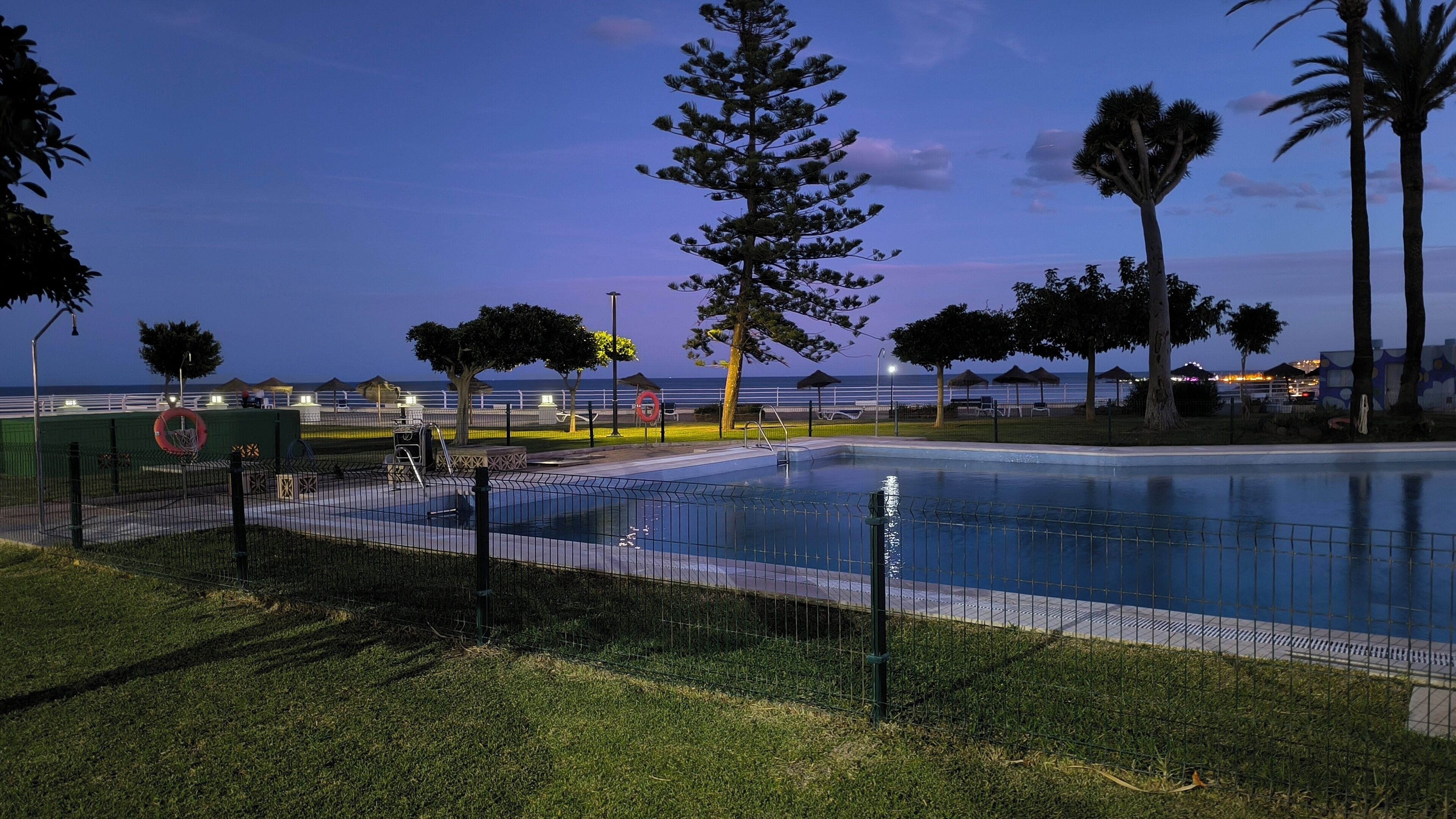 Una piscina con valla, rodeada de césped y árboles, con vista al mar y sombrillas de playa al fondo.