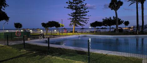 Una piscina con valla, rodeada de césped y árboles, con vista al mar y sombrillas de playa al fondo.