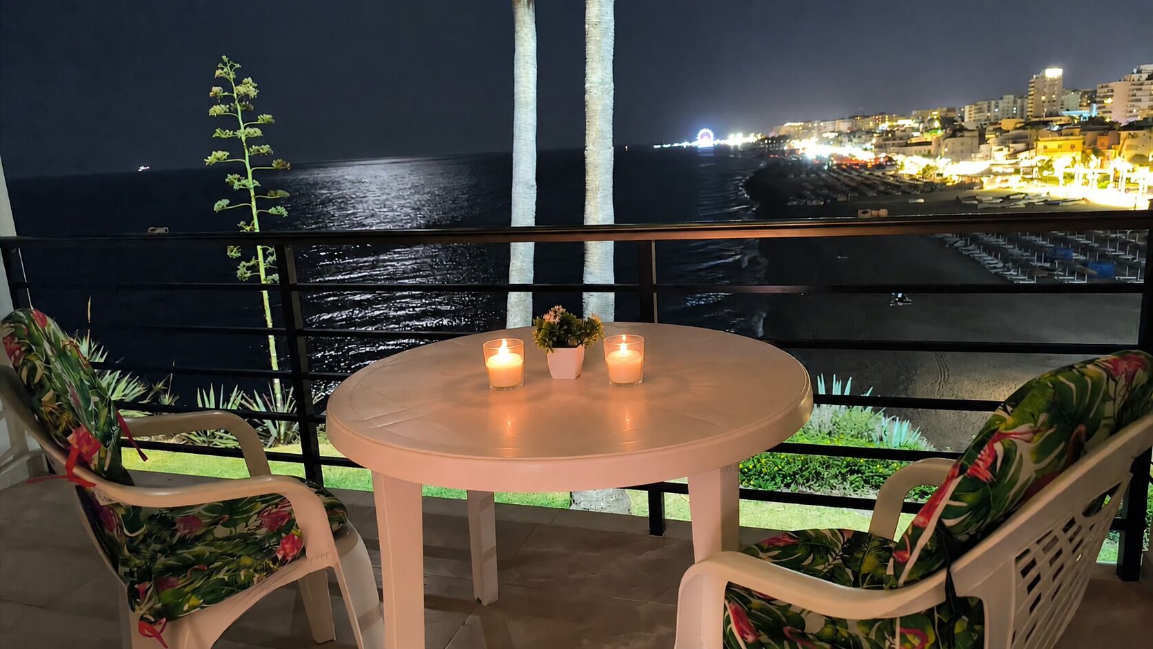 A balcony with a table and chairs, a view of the sea, and a cityscape at night.