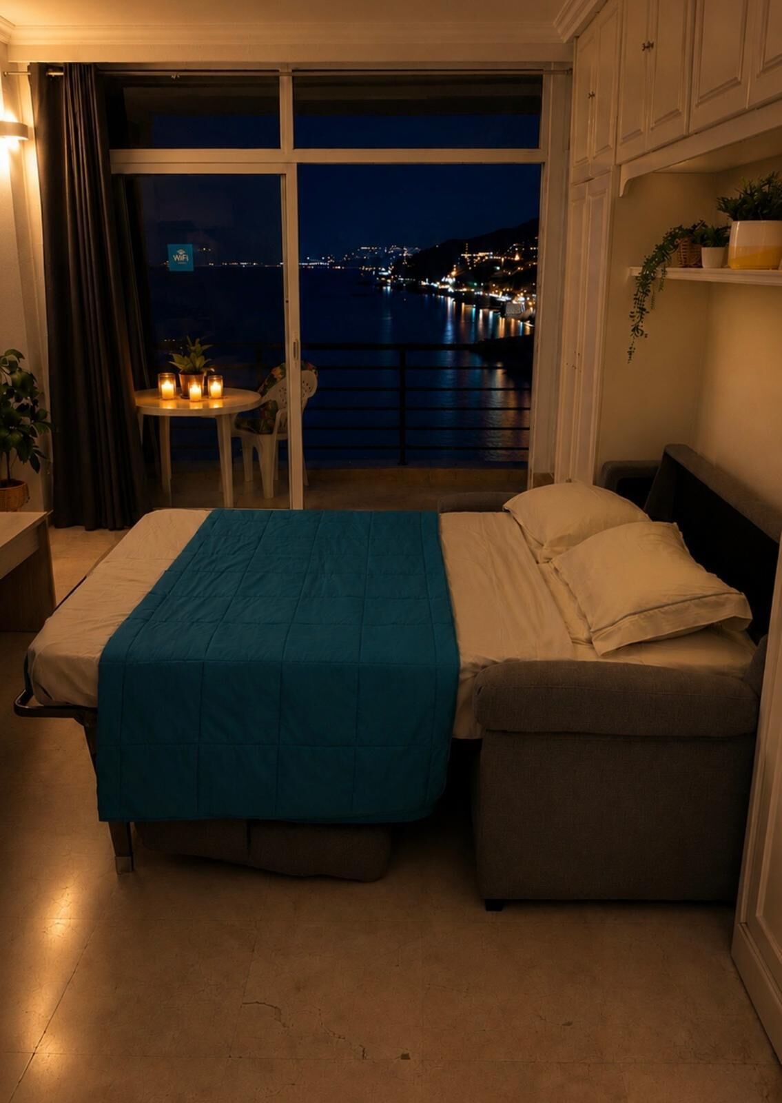 A bedroom with a bed, a small table with candles, a view of a cityscape at night, and a balcony with a railing.