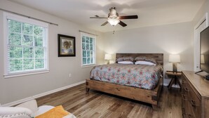A bedroom with a wooden bed, two bedside lamps, a ceiling fan, and large windows with blinds. - Beautiful Home Close To Everything (Greenville)