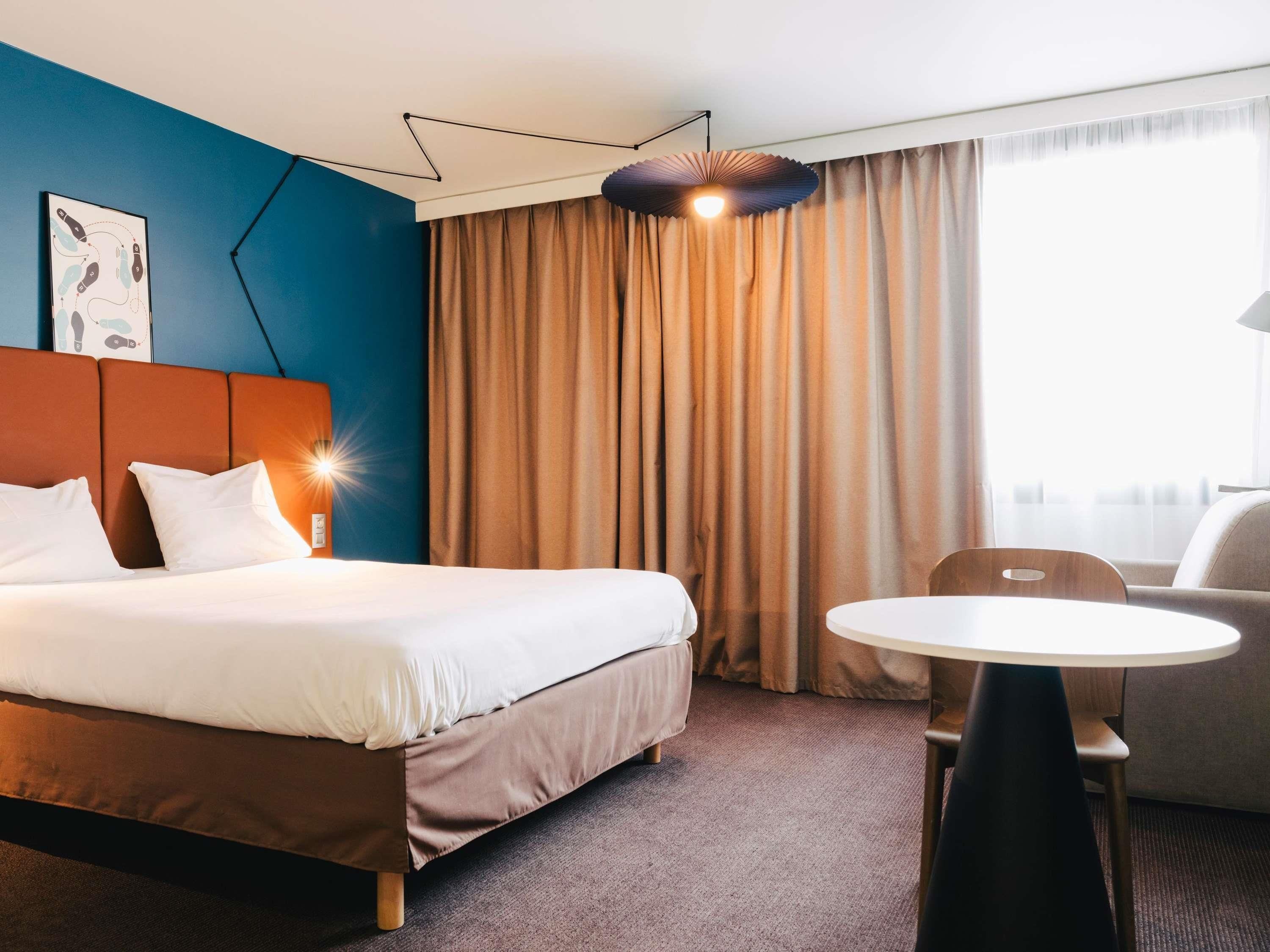 A hotel room with a bed, a desk, a chair, and a large window with curtains.