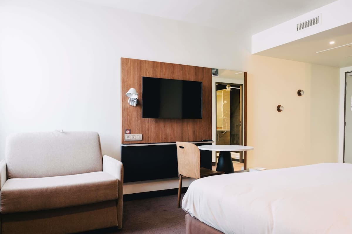 Ett hotellrum med en säng, ett skrivbord med en stol, en tv som är monterad på väggen och en dörr som leder till ett badrum.