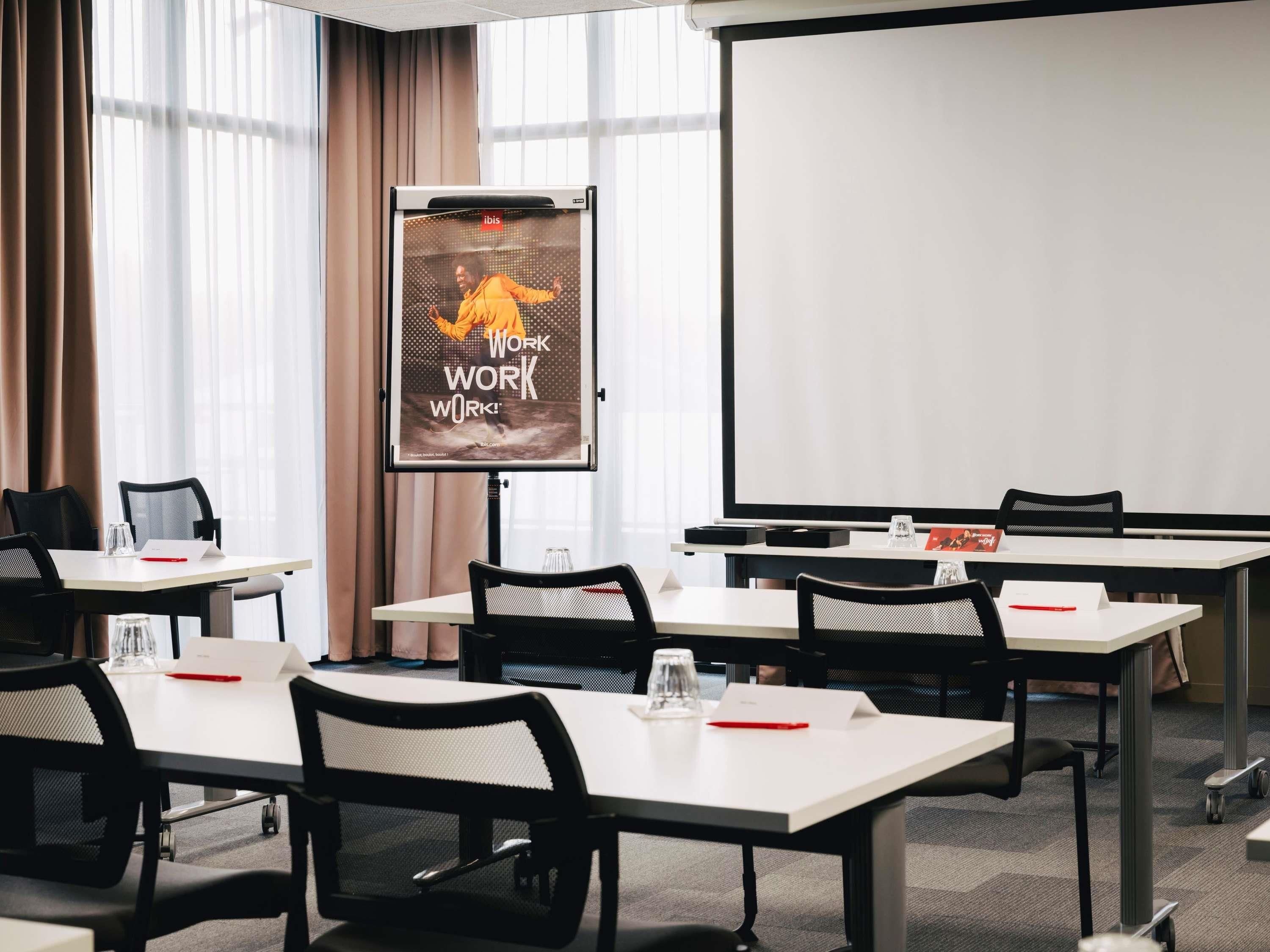Une salle de conférence équipée d’un écran de projection, d’un tableau blanc et de plusieurs tables avec des chaises.