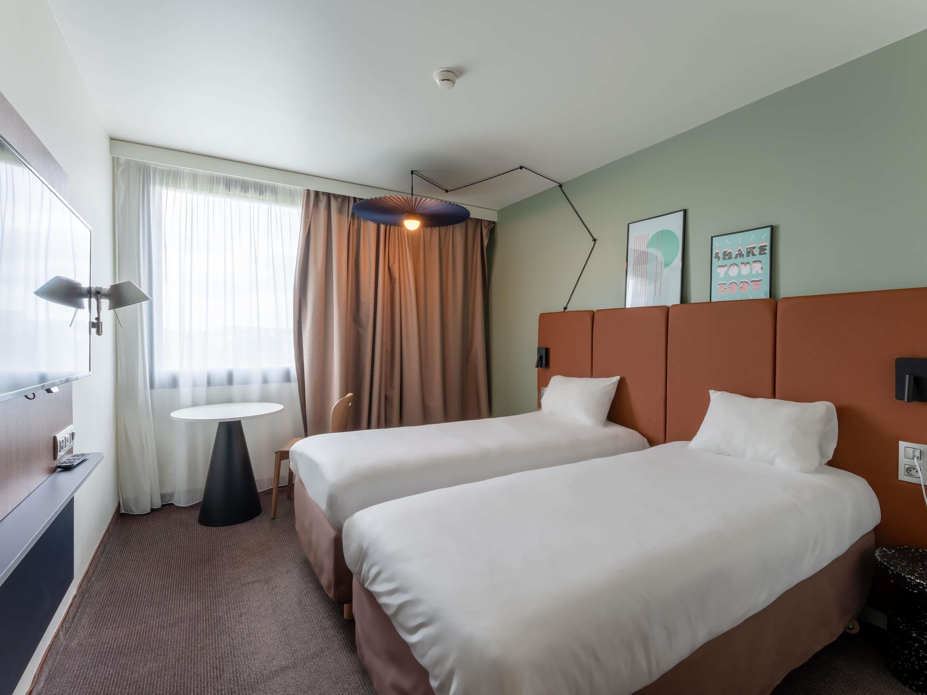 A hotel room with two beds, a desk, a chair, a small table, and a window with curtains.