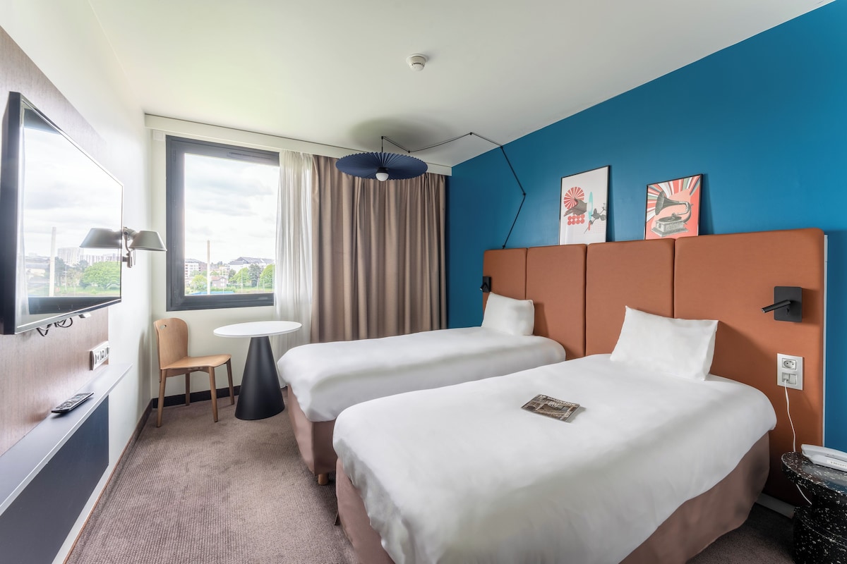 A hotel room with two beds, a TV, a small table, and a window with curtains.