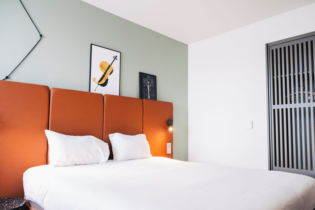 A hotel room with a large bed, orange headboard, white bedding, and two framed pictures on the wall.
